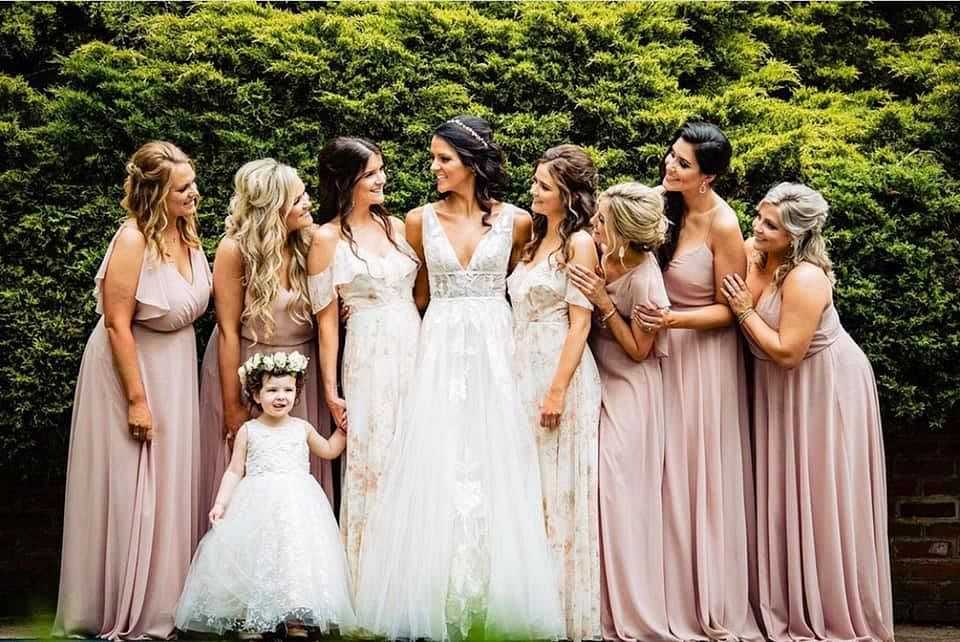 Bride and bridesmaids in pastel dresses pose with a flower girl in a garden setting.