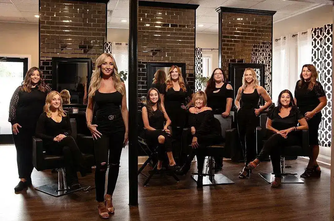 A team of nine women dressed in black poses in a hair salon with modern decor.
